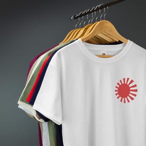 Op de afbeelding: Een wit T-shirt met een rode opkomende zon grafiek aan de rechterkant. Het shirt hangt aan een houten hanger met andere T-shirts in verschillende kleuren.