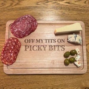 Könnte beinhalten: Ein rechteckiges Holz-Servierbrett mit geschnitztem Rand, das geschnittene Salami, Käse, Oliven und den eingravierten Text "OFF MY TITS ON PICKY BITS" zeigt. Das Brett liegt auf einer Holzoberfläche.