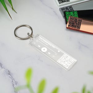 May include: A clear acrylic keychain with a music player design. It features a play button, rewind and fast forward buttons, and a QR code. The text "Your Custom Song Title" and "Your Custom Artist" is printed on the keychain.