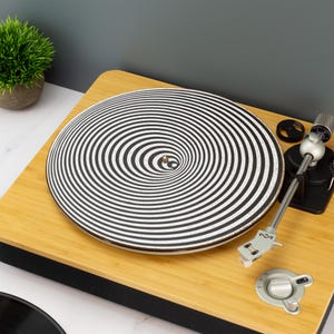 Może przedstawiać: Gramofon z jasnego drewna z płytą z czarno-białym spiralnym wzorem. Gramofon ma srebrne ramię i elementy sterujące. W tle widoczna jest mała roślina doniczkowa.