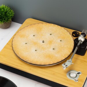 Może przedstawiać: Zbliżenie na gramofon z płytą na talerzu. Gramofon jest wykonany z drewna i ma czarną podstawę. Płyta jest jasnobrązowa i ma kilka małych otworów. Ramię gramofonu jest w dolnej pozycji i znajduje się nad płytą.