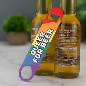 May include: A rainbow-colored bottle opener with the text "Queer for Beer" in black letters. The opener has a rainbow design with red, orange, yellow, green, blue, indigo, and violet stripes.