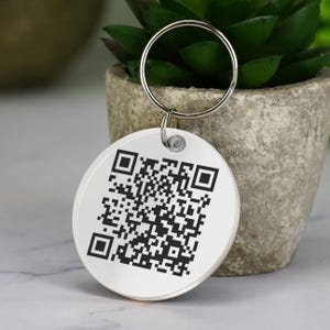 May include: A white round keyring with a black and white QR code printed on it. The keyring is attached to a silver key ring.
