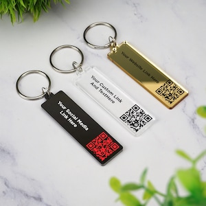 May include: Three rectangular keychains with silver key rings. One is black with red QR code and text "Your Social Media Link Here." Another is clear with a black QR code and text "Your Custom Link And Text Here." The third is gold with a black QR code and text "Your Website Link Here."