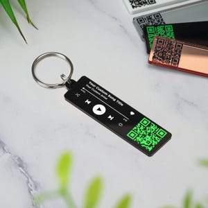May include: Black and green music player keychain with a QR code. The keychain has a play button, rewind button, and fast forward button. The text on the keychain reads "Your Custom Song Title" and "Your Custom Artist".