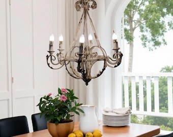 May include: An antique-style chandelier with six candle-style lights hangs above a wooden dining table. The chandelier has a distressed white and black finish. A potted plant with pink flowers, a white pitcher, and lemons are on the table.