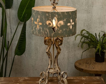Puede incluir: Una lámpara de mesa blanca desgastada con una base de metal decorativa y una pantalla de lámpara azul claro con diseños recortados. La pantalla de la lámpara tiene un diseño de flor de lis.