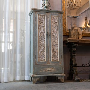 May include: A tall, gray-blue wooden cabinet with carved floral designs on the doors. The cabinet has a single drawer at the bottom and ornate hardware. The cabinet is a decorative storage piece.