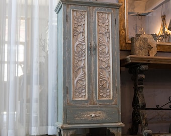 May include: A tall, gray-blue wooden cabinet with carved floral designs on the doors. The cabinet has a single drawer at the bottom and ornate hardware. The cabinet is a decorative storage piece.