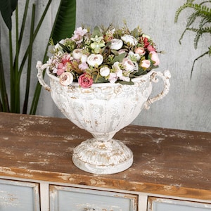 May include: A distressed white urn-shaped planter filled with an assortment of artificial flowers in shades of pink, white, and green. The planter has ornate handles and sits on a pedestal base. The arrangement is on a wooden surface.