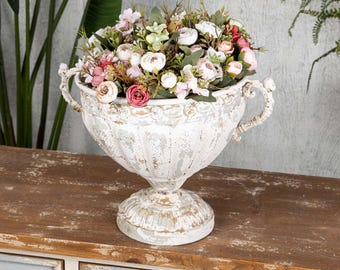 Op de afbeelding: Een witte, verweerde urnevormige plantenbak gevuld met kunstbloemen in roze, wit en groen. De plantenbak heeft sierlijke handvatten en staat op een voetstuk. De opstelling staat op een houten oppervlak.