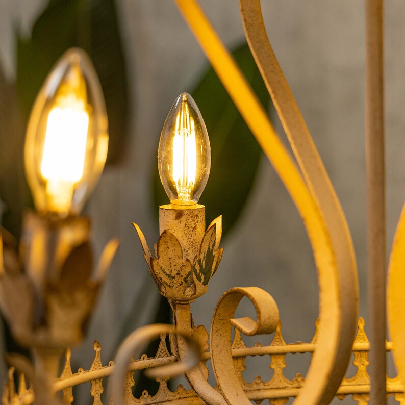 May include: A close-up of a vintage-style chandelier with two lit light bulbs. The chandelier is made of metal and has a distressed white finish. The light bulbs are clear glass with a filament design.