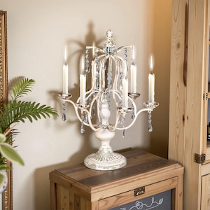 May include: A white metal candelabra with a distressed finish and crystal accents. The candelabra has six arms and holds six candles. It is sitting on a wooden table.