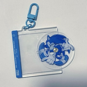 May include: A clear acrylic keychain featuring a blue cartoon character, Sonic the Hedgehog, with a blue carabiner clip. The keychain has a clear plastic holder with text that reads "SONIC ADVENTURE 2 / ORIGINAL SOUNDTRACK".