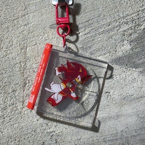 May include: A clear acrylic keychain featuring a red and black character from the Sonic the Hedgehog series. The keychain is attached to a red metal clasp. A red label with white text is also attached.