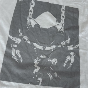 May include: A white t-shirt featuring a large, dark gray rectangular graphic print. The design depicts a stylized, pixelated illustration of a panther head with a chain-link necklace draped around its neck, rendered in high-contrast white lines.