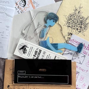 May include: A clear acrylic standee of a blue-toned character sitting down, placed over a Faraway Town Youth Ensemble Fall Recital ticket and a black rectangular display piece with the text "??? Remember to be patient..." printed in white.