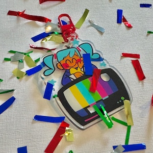May include: A clear acrylic keyring featuring a cartoon character with blue hair and a colourful TV screen design. The keyring has a red clasp and is surrounded by colourful confetti.