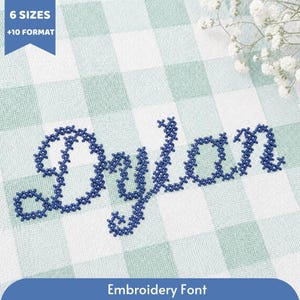 May include: A dark blue cross-stitch embroidery design of the name Dylan in a cursive script font. The text is displayed on a light green and white gingham fabric background. A blue banner in the top left corner contains the text 6 SIZES +10 FORMAT.