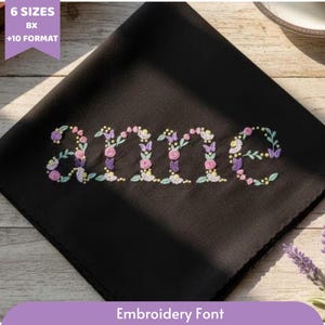 Könnte beinhalten: Schwarzer quadratischer Stoff mit dem Namen "anne", der in einer floralen Schriftart gestickt ist. Die Buchstaben sind mit rosa, lila und gelben Blumen und Blättern verziert. Der Text "Embroidery Font" steht unten.