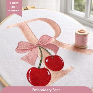 Puede incluir: Diseño bordado con una letra "R" rosa, un lazo y cerezas rojas sobre tela blanca, en un bastidor de madera. Una bobina de hilo rosa está al lado. El texto dice "Embroidery Font".