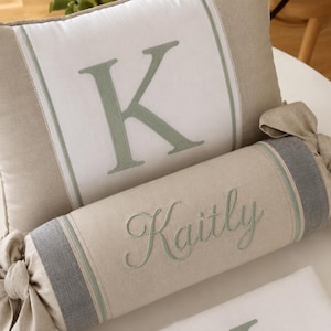 May include: A rectangular throw pillow with a white center panel featuring a large embroidered sage green letter K, flanked by beige sides. Below it, a cylindrical bolster pillow in beige and gray features the embroidered name Kaitly.