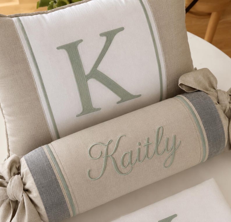 May include: A rectangular throw pillow with a white center panel featuring a large embroidered sage green letter K, flanked by beige sides. Below it, a cylindrical bolster pillow in beige and gray features the embroidered name Kaitly.