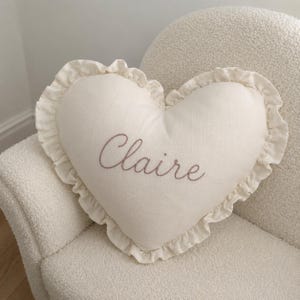 May include: A heart-shaped off-white throw pillow featuring a ruffled edge and the name Claire embroidered in a brown cursive script across the center. The pillow is made of a textured fabric and maintains a consistent color palette throughout.