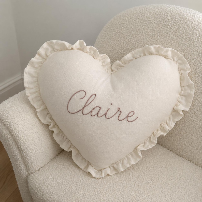 May include: A heart-shaped off-white throw pillow featuring a ruffled edge and the name Claire embroidered in a brown cursive script across the center. The pillow is made of a textured fabric and maintains a consistent color palette throughout.