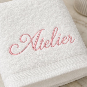 May include: A white terry cloth towel featuring the word Atelier embroidered in a pink cursive script font. The towel has a textured surface and a decorative woven border along the bottom edge, providing a clean and simple aesthetic.