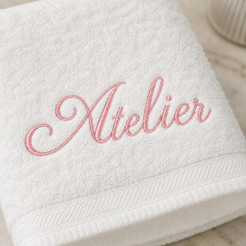 May include: A white terry cloth towel featuring the word Atelier embroidered in a pink cursive script font. The towel has a textured surface and a decorative woven border along the bottom edge, providing a clean and simple aesthetic.