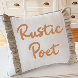 May include: Square white cushion embroidered with the words "Rustic Poet" in orange script. It features a ruffled border in a neutral beige colour, adding a touch of rustic charm to the design.