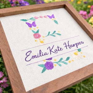 May include: A framed embroidered artwork. The design includes the name "Emilia Kate Harper" in purple script, encircled by floral and butterfly embroidery in pink, purple, yellow, and green. The background is a light-coloured fabric, set within a wooden frame.