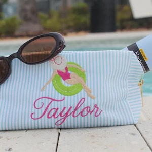 May include: A light blue and white striped zippered pouch featuring an embroidered design of a figure in a pink swimsuit and white hat floating in a green inner tube, with the name Taylor embroidered in pink script and black sunglasses.