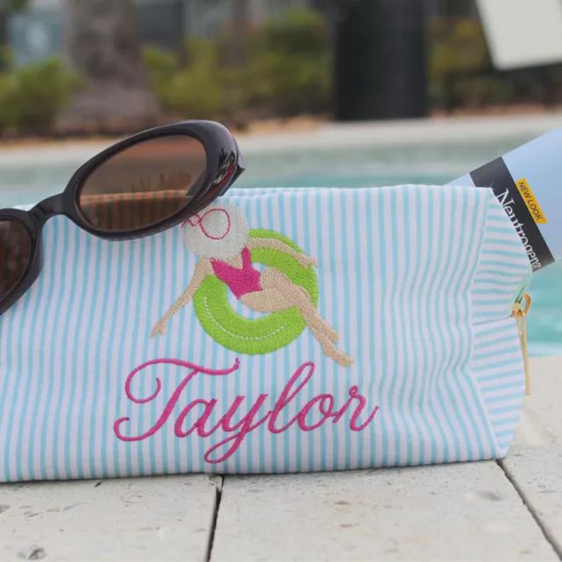 May include: A light blue and white striped zippered pouch featuring an embroidered design of a figure in a pink swimsuit and white hat floating in a green inner tube, with the name Taylor embroidered in pink script and black sunglasses.