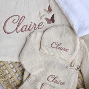 May include: A cream-colored knitted baby set including a long-sleeve wrap top, matching beanie, and a muslin blanket. The name Claire is embroidered in dusty rose thread on each piece, accompanied by three small butterfly motifs on the blanket.