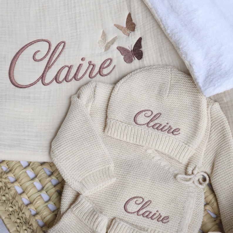 May include: A cream-colored knitted baby set including a long-sleeve wrap top, matching beanie, and a muslin blanket. The name Claire is embroidered in dusty rose thread on each piece, accompanied by three small butterfly motifs on the blanket.