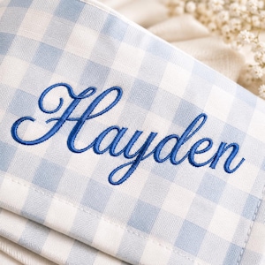 May include: A light blue and white gingham fabric item featuring the name Hayden embroidered in a dark blue cursive script font. The textile has a woven checkered pattern with visible stitching along the edges and a soft, folded appearance.