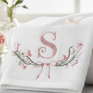 May include: White bath towel with an embroidered pink letter "S" and floral design. The embroidery includes pink bell-shaped flowers, green stems, and a pink bow. The towel is folded and appears soft.