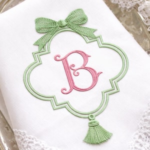 Op de afbeelding: Witte linnen servet met een groene strik en een decoratief groen geborduurd frame. Binnen het frame staat de roze letter "B" en een groene kwast. De servet heeft een kanten rand.