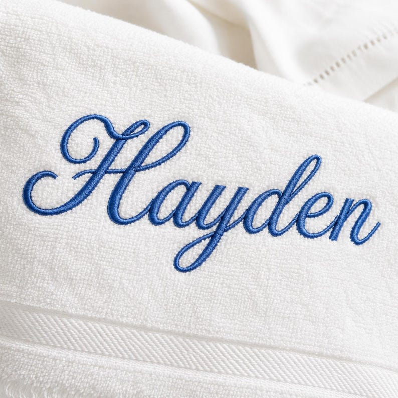 May include: A white terry cloth towel featuring the name Hayden embroidered in a blue cursive script font. The towel has a textured surface with a horizontal decorative border detail visible near the bottom edge of the fabric.