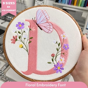 Könnte beinhalten: Gesticktes Blumendesign mit einem rosa Buchstaben "D", einem Schmetterling und Blumen in einem Holzreifen. Die Stickerei umfasst lila, rosa, gelbe und weiße Blumen. Der Text "Floral Embroidery Font" steht unten.