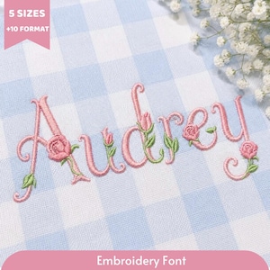 May include: A light blue and white gingham fabric featuring the embroidered name Audrey in pink cursive script. The letters are decorated with small pink rosebuds and green leaves. A pink banner in the corner reads 5 SIZES +10 FORMAT.