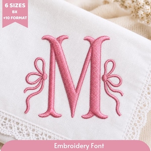 May include: A white fabric featuring a pink embroidered letter M flanked by two decorative bows. The text 6 SIZES BX +10 FORMAT appears in a pink banner at the top left, and the text Embroidery Font appears in a pink banner at the bottom.
