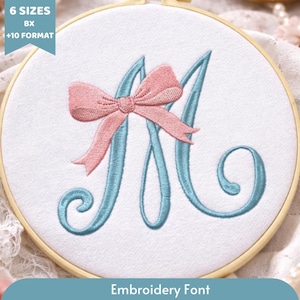 May include: An embroidery hoop featuring a light blue letter "M" with a pink bow. The text "6 SIZES", "BX", and "+10 FORMAT" is in the upper left corner. The text "Embroidery Font" is at the bottom.