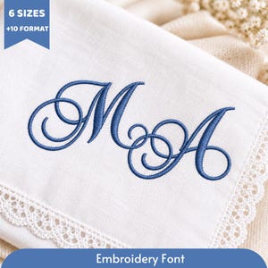 Puede incluir: Una pieza de tela blanca con las letras cursivas M y A bordadas en azul. Un estandarte azul en la esquina superior izquierda muestra el texto 6 SIZES +10 FORMAT. Un estandarte azul en la parte inferior contiene el texto Embroidery Font.