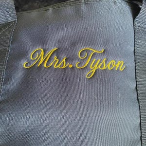 May include: A dark gray duffel bag featuring the embroidered text Mrs. Tyson in a gold cursive script. The bag is constructed from a durable, woven fabric with thick, matching gray nylon straps attached to the sides of the main compartment.