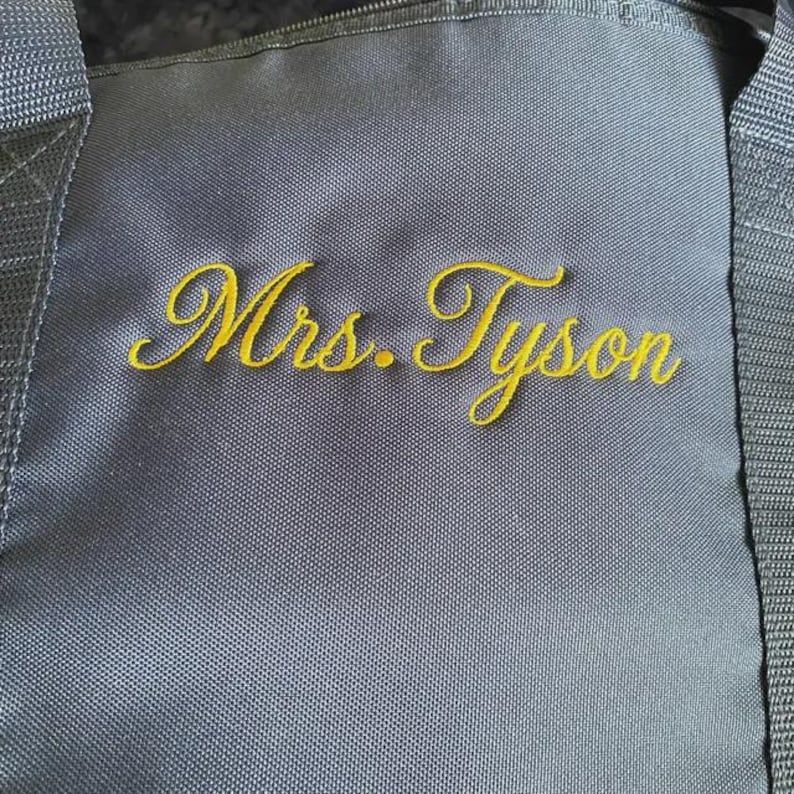 May include: A dark gray duffel bag featuring the embroidered text Mrs. Tyson in a gold cursive script. The bag is constructed from a durable, woven fabric with thick, matching gray nylon straps attached to the sides of the main compartment.