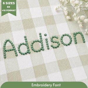 Può includere: Un design di carattere per ricamo che mostra il nome addison in lettere minuscole verdi formate da piccoli punti circolari. Il testo è posizionato su uno sfondo di tessuto a quadri verde chiaro e bianco con fiori bianchi nell'angolo.