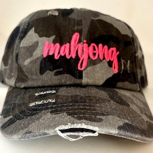 May include: A distressed camouflage baseball cap in shades of gray and black. The word "mahjong" is embroidered in bright pink cursive across the front. The cap has a pre-curved bill and distressed detailing.